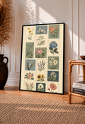 Patchwork of flower and leaf stamp style panels in sage blue pink and cream in a black frame leaning on a sunlit wooden floor beside books and a rattan seat