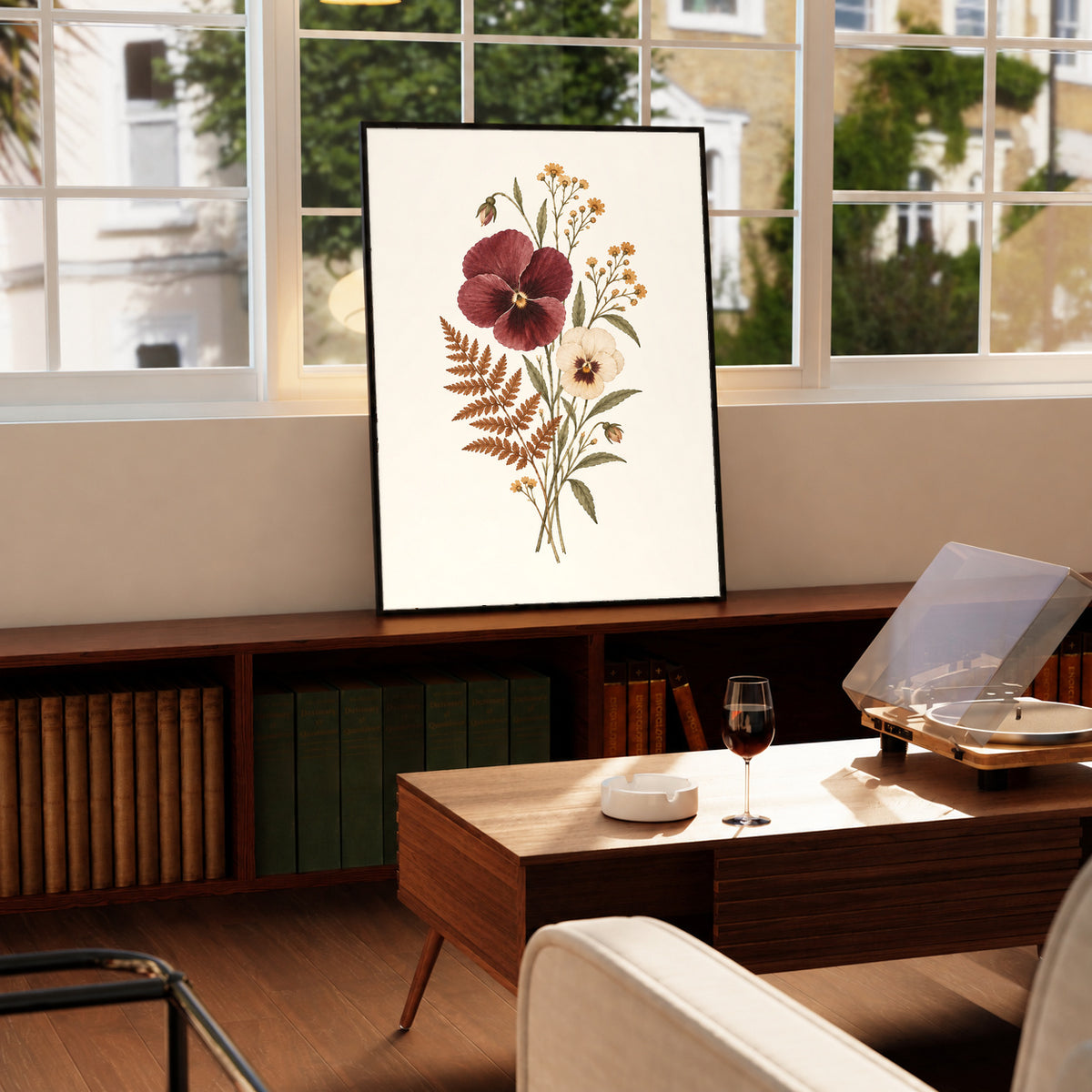 Burgundy and cream pansies with rust fern leaves and small golden flowers in a black frame on a wooden cabinet beside a window and record player