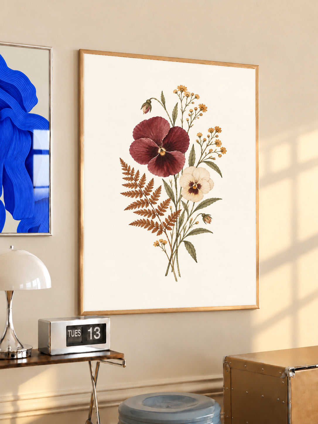 Burgundy and cream pansies with rust fern leaves and small golden flowers in an oak frame above a console beside a blue print and calendar