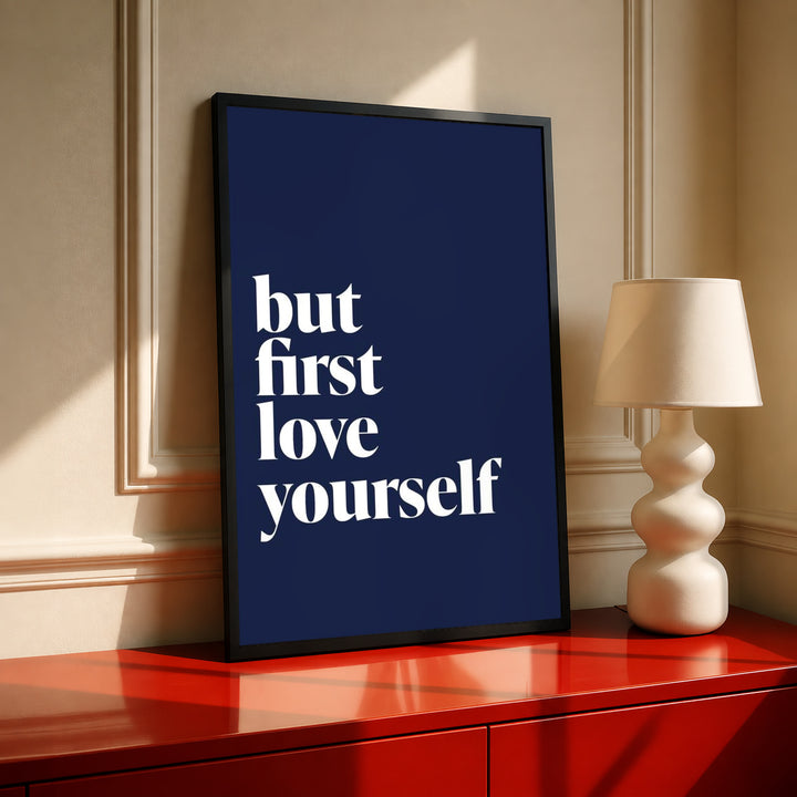 Navy blue poster in dark frame with white serif text "but first love yourself"