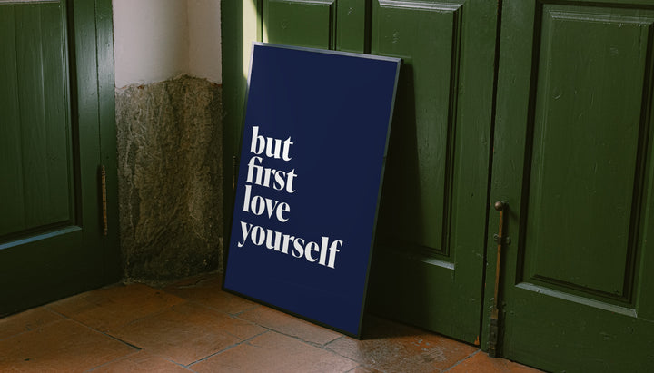 Navy blue framed poster leaning with white serif quote "but first love yourself"