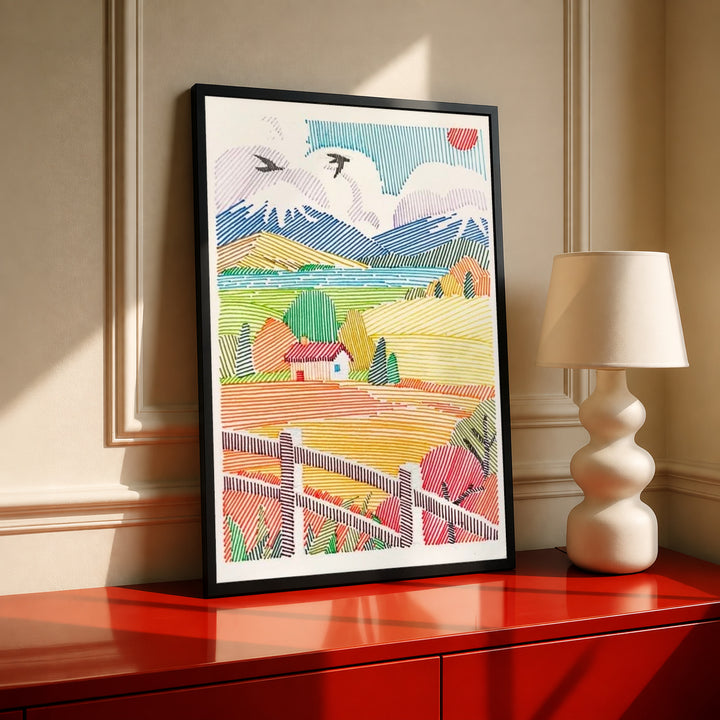A red-roof farmhouse among colorful striped fields, mountains, birds and a foreground fence in a black frame on a red console beside a sculptural cream lamp
