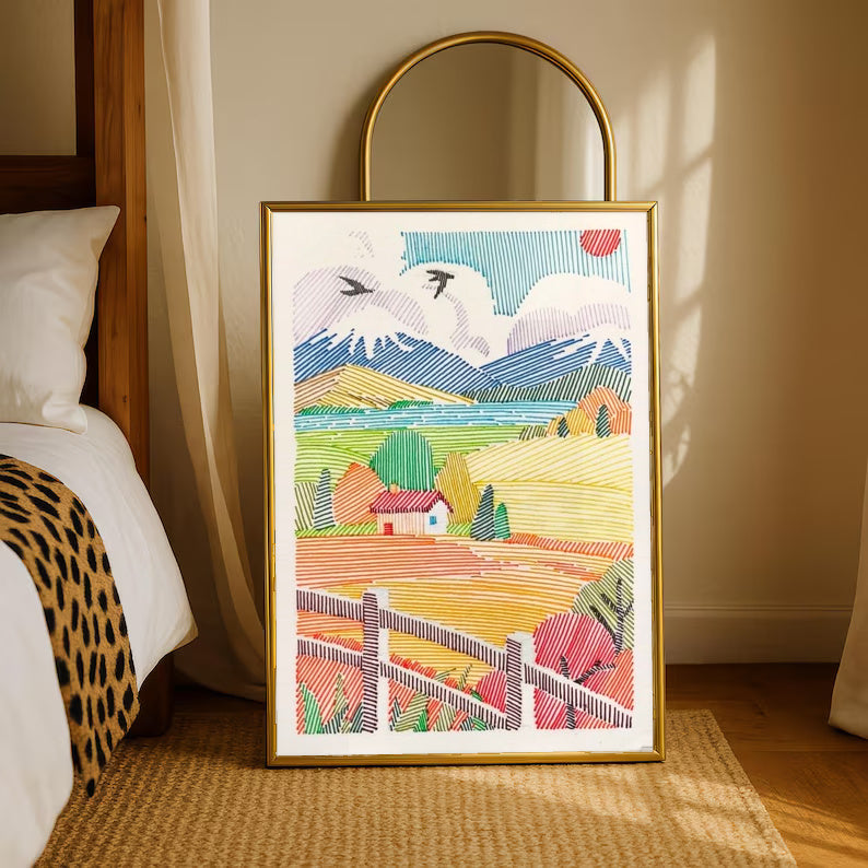 A red-roof farmhouse among colorful striped fields, mountains, birds and a foreground fence in a gold-tone styling frame leaning beside a bedroom mirror