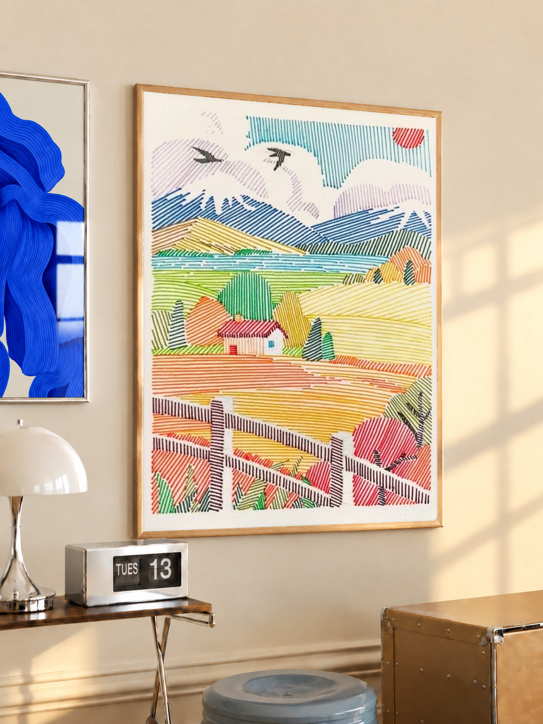 A red-roof farmhouse among colorful striped fields, mountains, birds and a foreground fence in a light wood frame on a sunlit office wall beside a desk