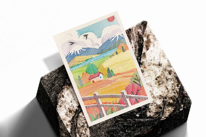 A red-roof farmhouse among colorful striped fields, mountains, birds and a foreground fence as an unframed print resting at an angle on a dark stone display