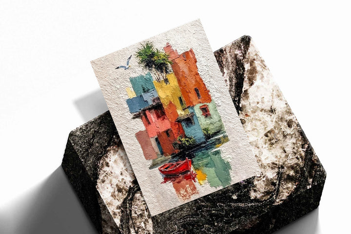Colorful waterfront houses reflected in green water beside a small red boat as an unframed print resting at an angle on a dark stone display