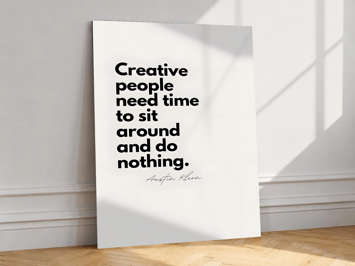 Creative people need time to sit around and do nothing quote in bold black type on cream, shown unframed on a white wall above parquet flooring with a diagonal sun shadow