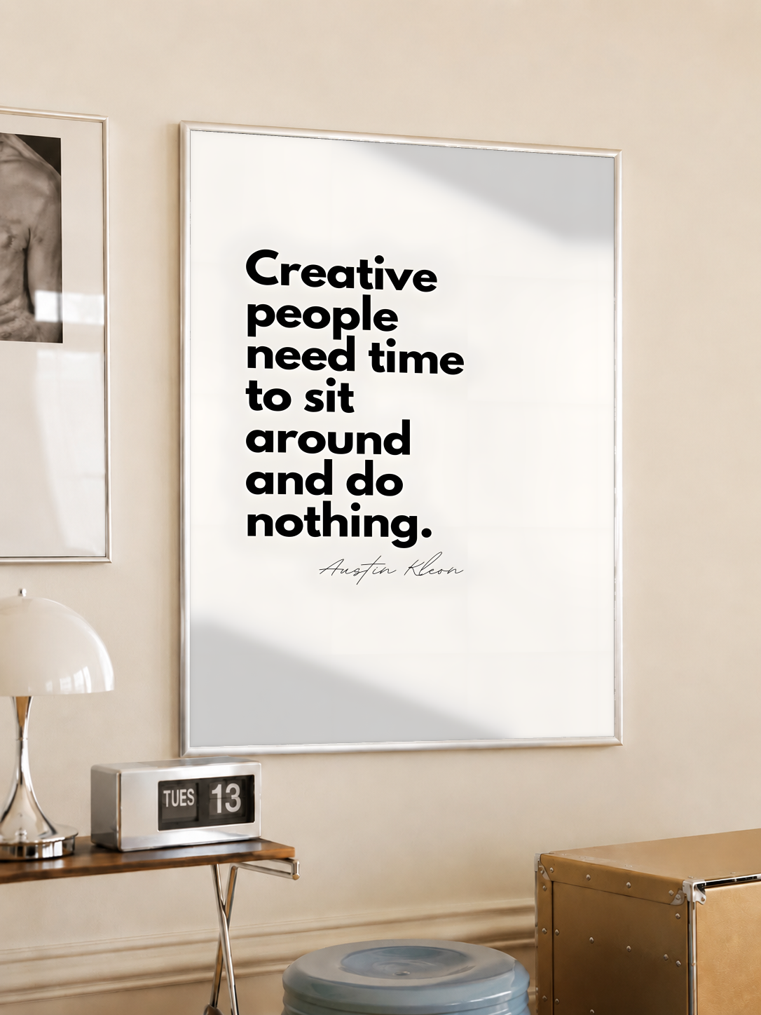 Creative people need time to sit around and do nothing quote in bold black type on cream, styled in a light frame on a cream wall beside a white dome lamp and glass side table