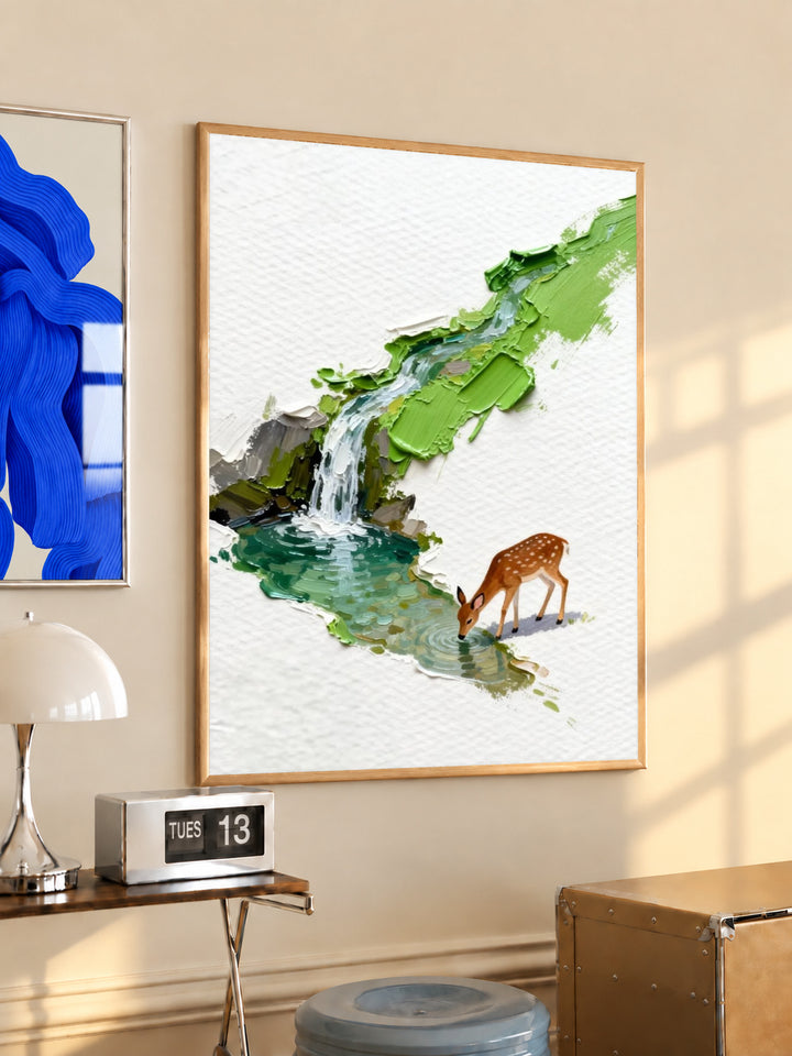 A small deer drinking beside a turquoise stream and waterfall under bright green foliage in a light wood frame on a sunlit office wall beside a desk