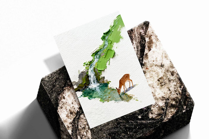 A small deer drinking beside a turquoise stream and waterfall under bright green foliage as an unframed print resting at an angle on a dark stone display