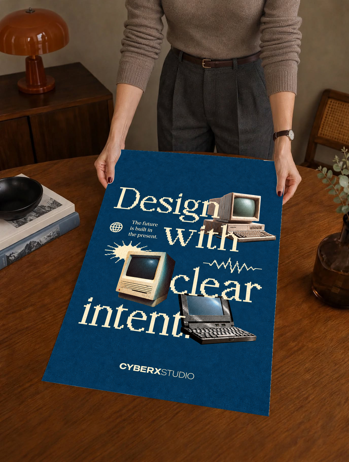 Vintage desktop computers and a laptop with cream pixel-style typography on blue as a large unframed poster held above a wooden desk