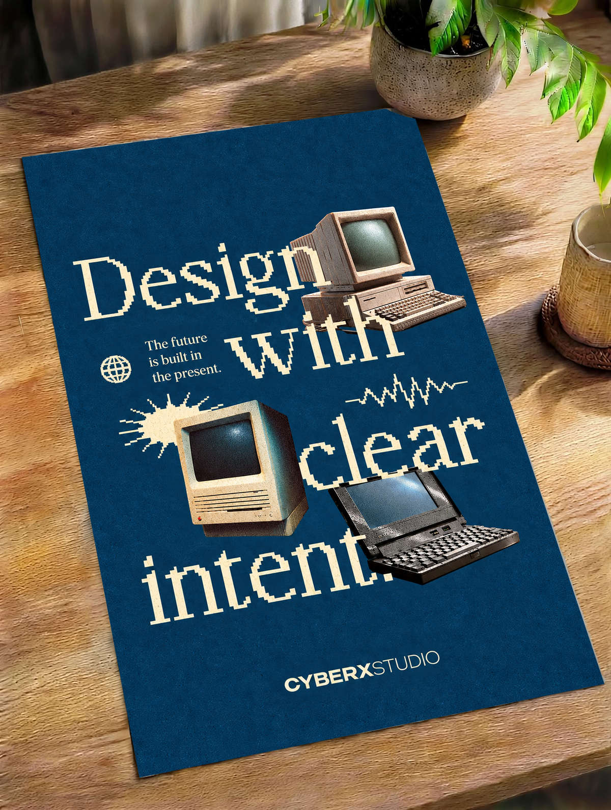 Vintage desktop computers and a laptop with cream pixel-style typography on blue as an unframed poster lying on a wooden table beside a plant