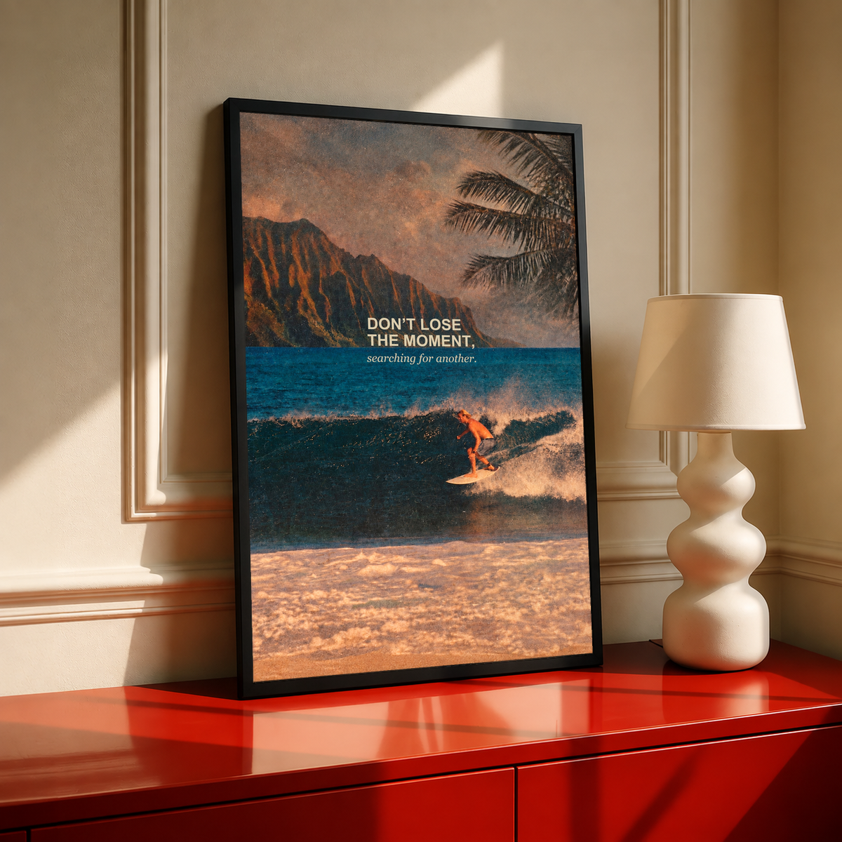 Surfer on a teal wave below warm sunset sky and palm fronds; black framed print on a red cabinet