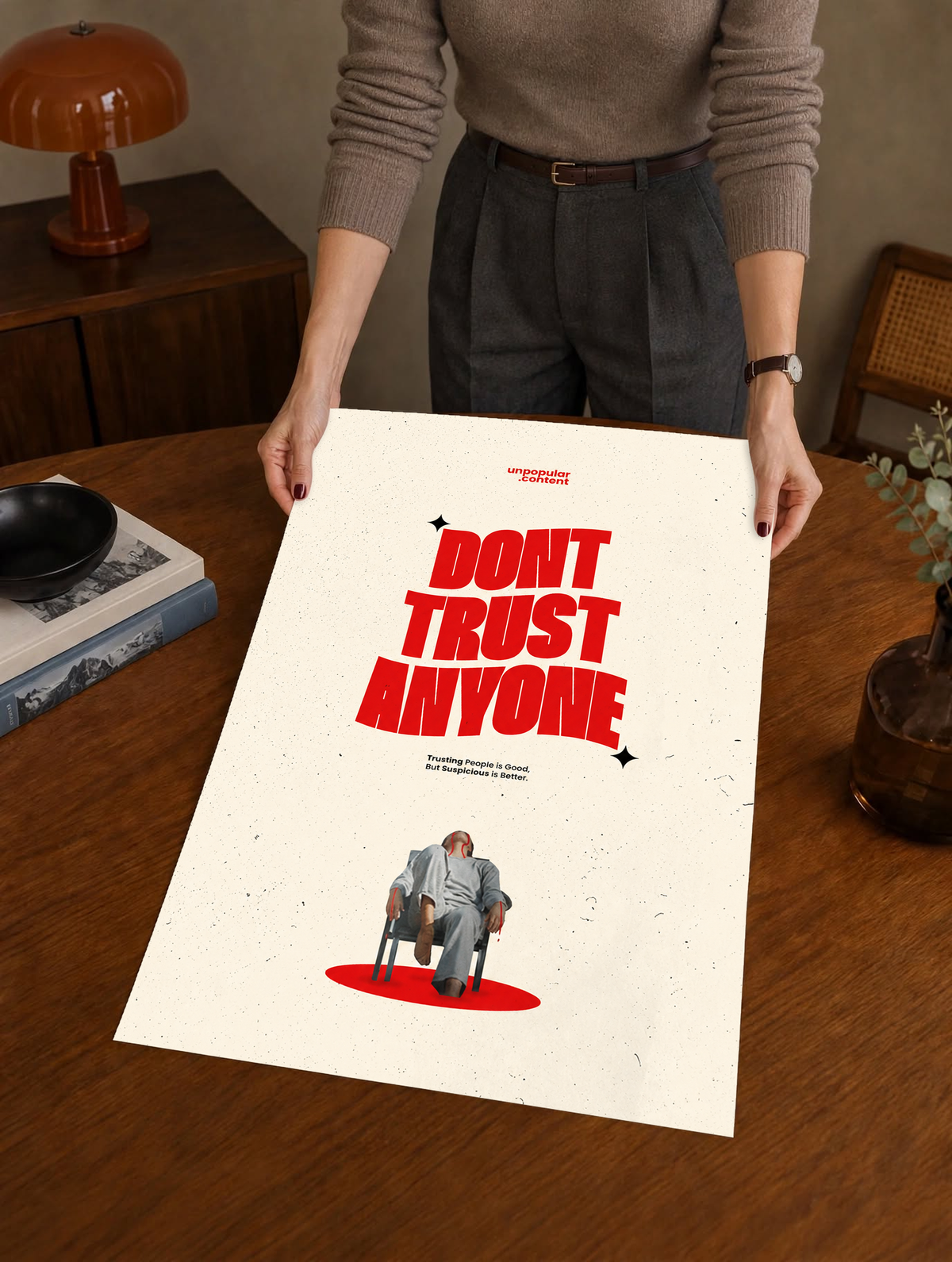 Bold red Don't Trust Anyone typography above a small seated figure on cream as a large unframed poster held above a wooden desk