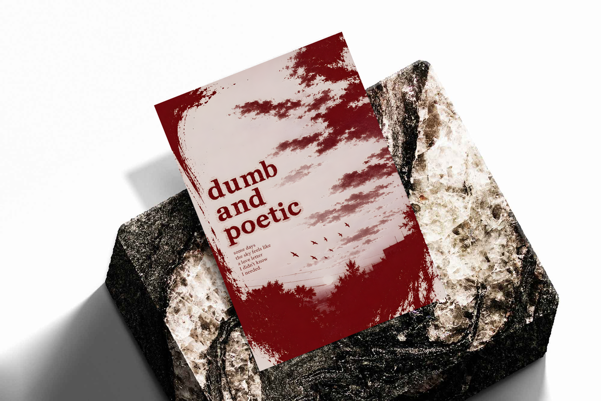 Dumb and poetic burgundy distressed lettering over pale pink clouds with birds and tree silhouettes, shown as an unframed print tilted on a dark stone display against a light background