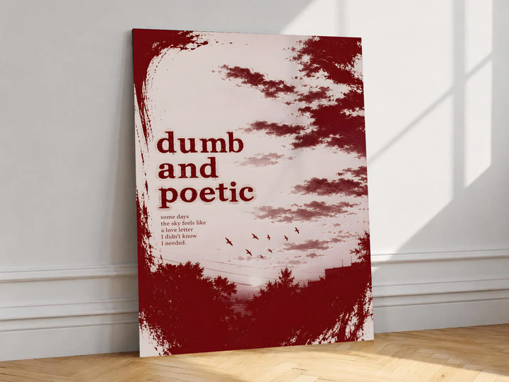 Dumb and poetic burgundy distressed lettering over pale pink clouds with birds and tree silhouettes, shown unframed on a white wall above parquet flooring with a diagonal sun shadow