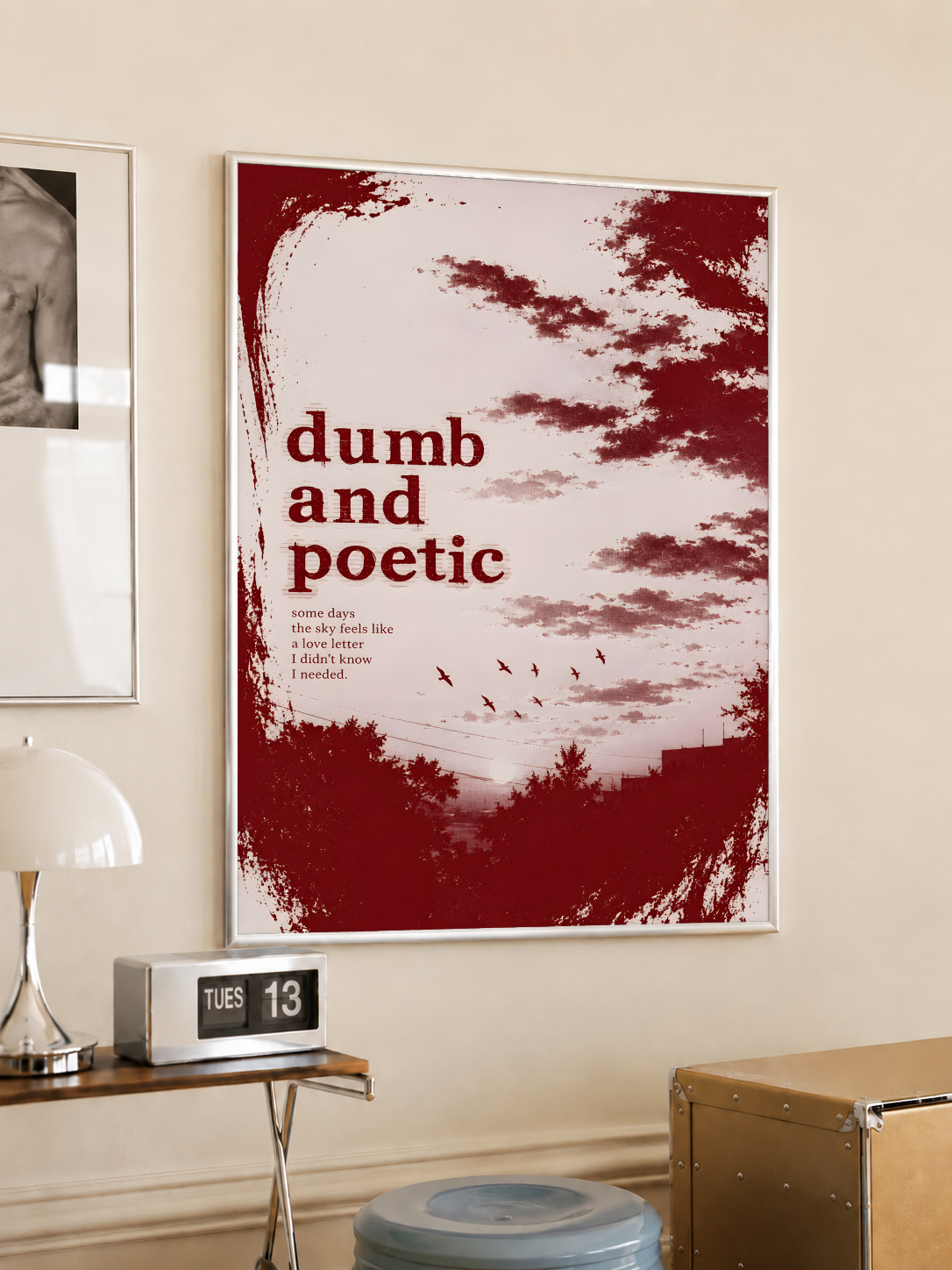 Dumb and poetic burgundy distressed lettering over pale pink clouds with birds and tree silhouettes, styled in a light frame on a cream wall beside a white dome lamp and glass side table