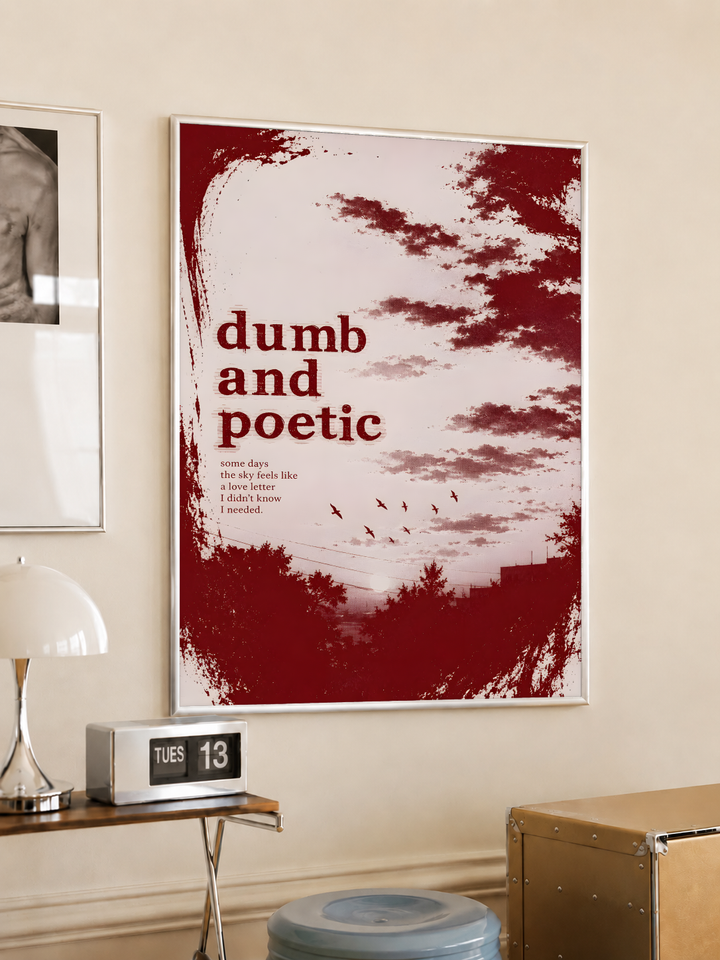 Dumb and poetic burgundy distressed lettering over pale pink clouds with birds and tree silhouettes, styled in a light frame on a cream wall beside a white dome lamp and glass side table