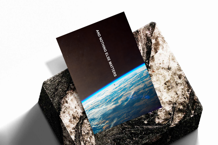 And Nothing Else Matters white vertical typography over Earth’s blue atmospheric horizon and a tiny human-shaped silhouette, shown as an unframed print tilted on a dark stone display against a light background