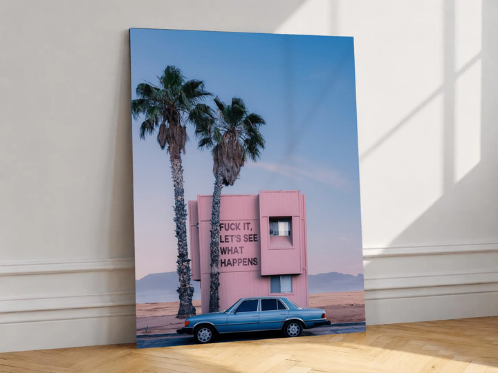 Canvas artwork depicting pink desert building, palms, blue classic car and irreverent quote, leaning against a white wall in angled sunlight.