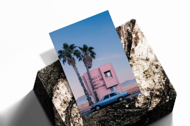 Unframed artwork depicting pink desert building, palms, blue classic car and irreverent quote, presented diagonally on rough stone.
