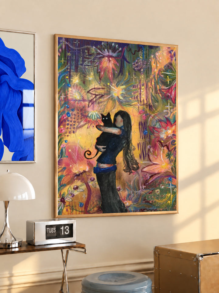 A girl embracing a black cat amid colorful ornamental flowers and patterns in a light wood frame on a sunlit office wall beside a desk