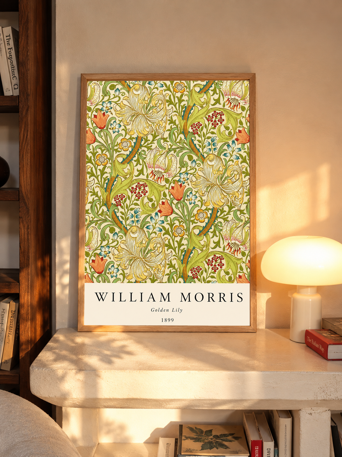 Golden Lily floral pattern with olive leaves, yellow flowers and red accents in an oak frame on a sunlit console beside books and a cream lamp