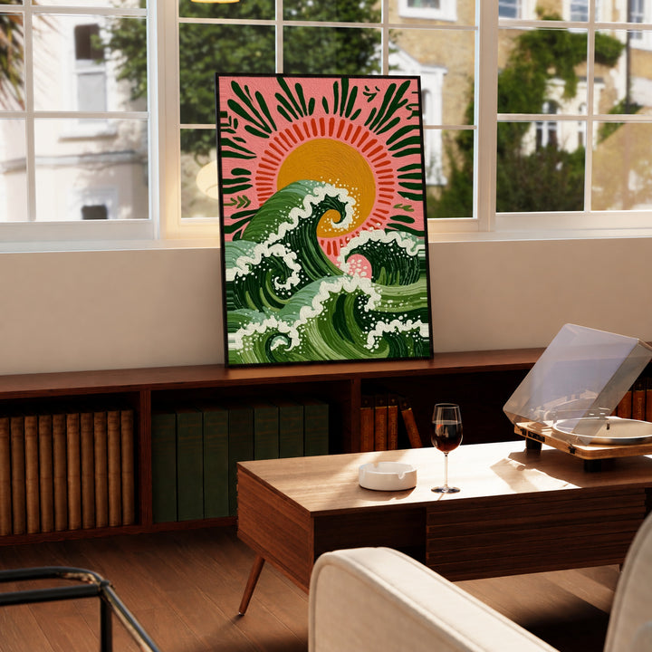 Curling green waves with cream foam beneath an orange sun on a pink background in a black display frame on a wooden console beside a sunlit window