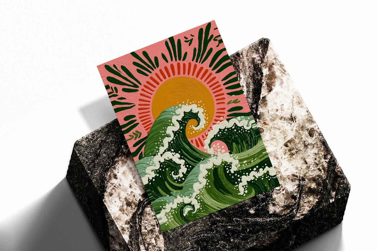Curling green waves with cream foam beneath an orange sun on a pink background as an unframed print resting at an angle on a dark stone display