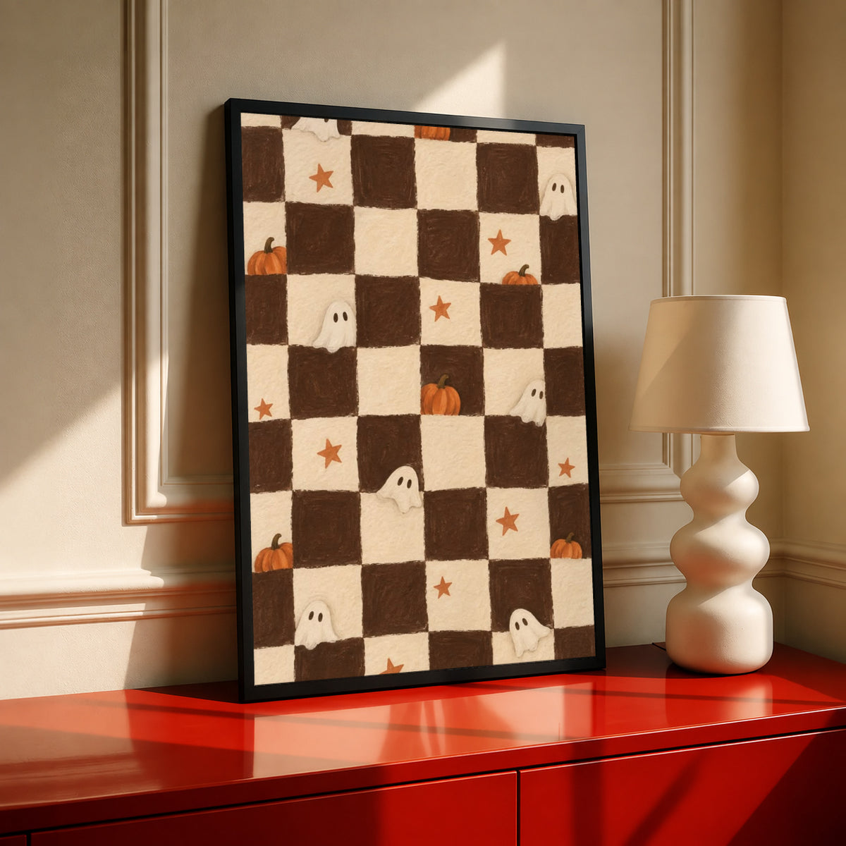 Brown and cream Halloween checkerboard with little white ghosts, pumpkins and orange stars in a black frame on a red console beside a sculptural cream lamp