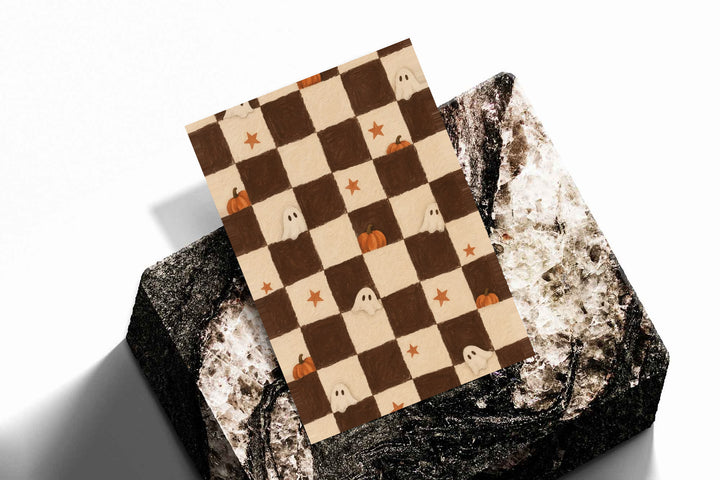 Brown and cream Halloween checkerboard with little white ghosts, pumpkins and orange stars as an unframed print resting at an angle on a dark stone display