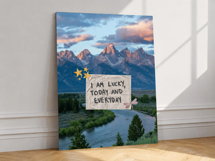 Canvas artwork depicting mountain and river scene behind a lucky affirmation note, leaning against a white wall in angled sunlight.
