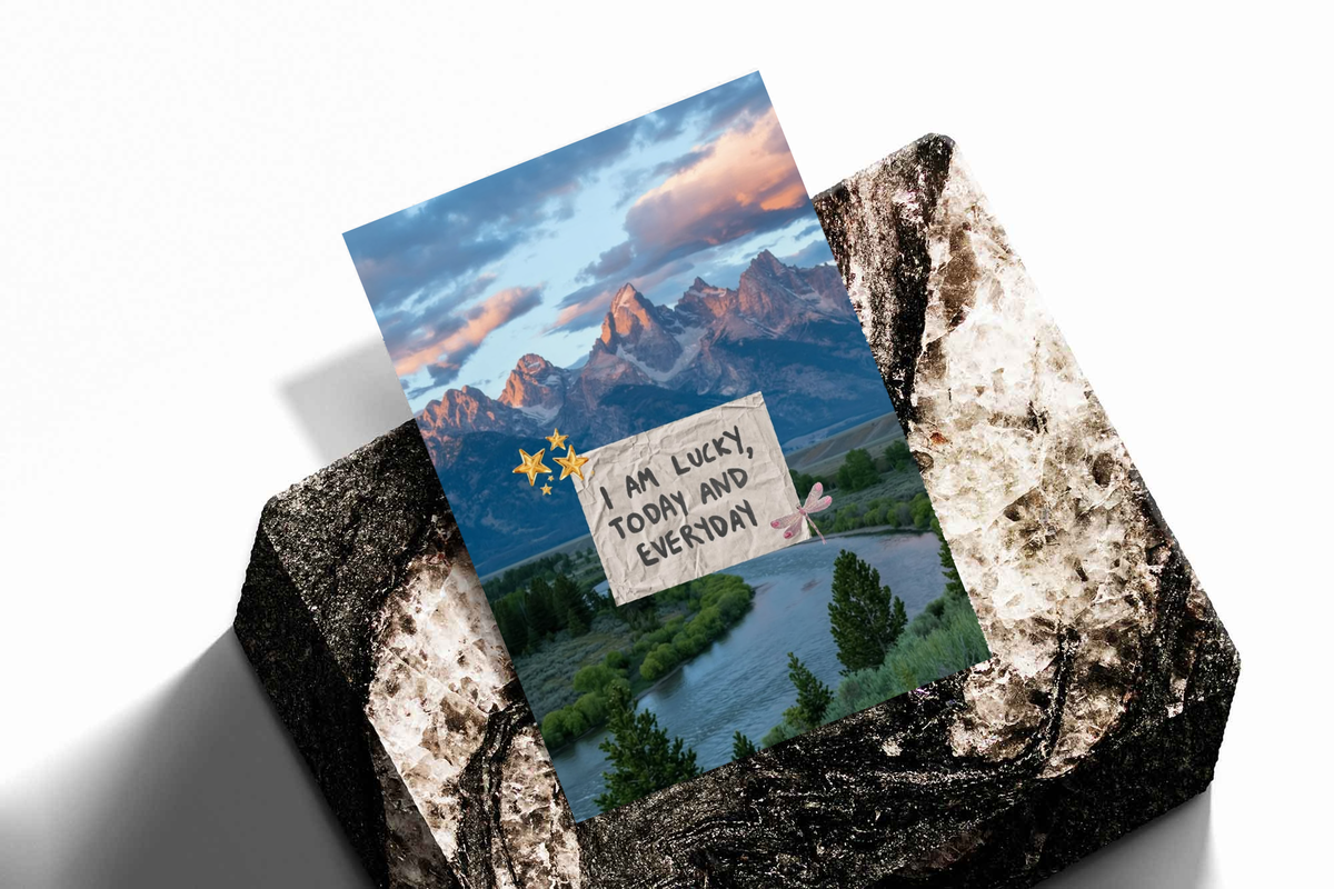 Unframed artwork depicting mountain and river scene behind a lucky affirmation note, presented diagonally on rough stone.