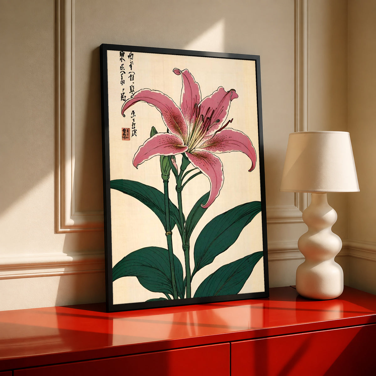 Pink lily with dark green leaves Japanese calligraphy and a red seal on cream in a black frame on a red cabinet beside a cream lamp