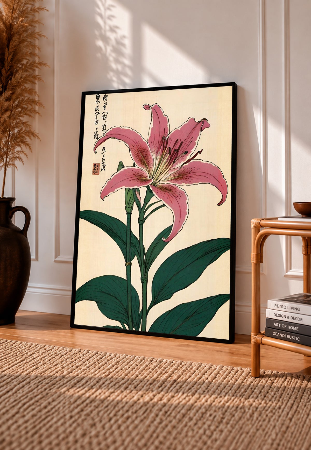 Pink lily with dark green leaves Japanese calligraphy and a red seal on cream in a black frame leaning on a sunlit wooden floor beside books and a rattan seat