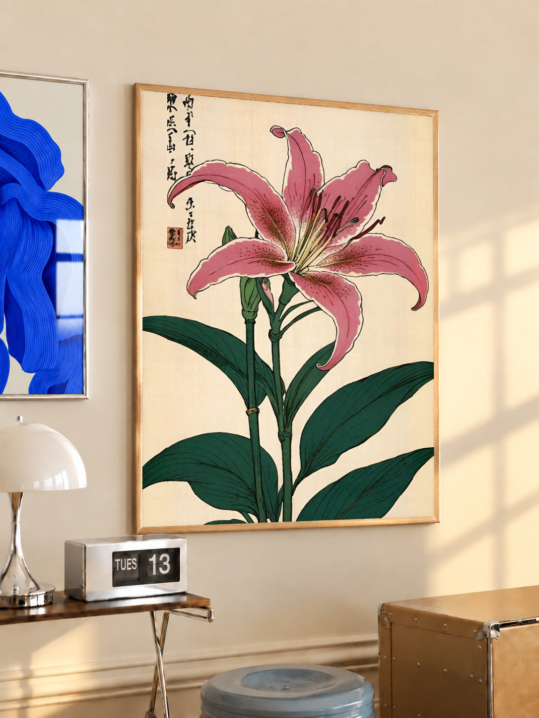 Pink lily with dark green leaves Japanese calligraphy and a red seal on cream in an oak frame above a console beside a blue print and calendar
