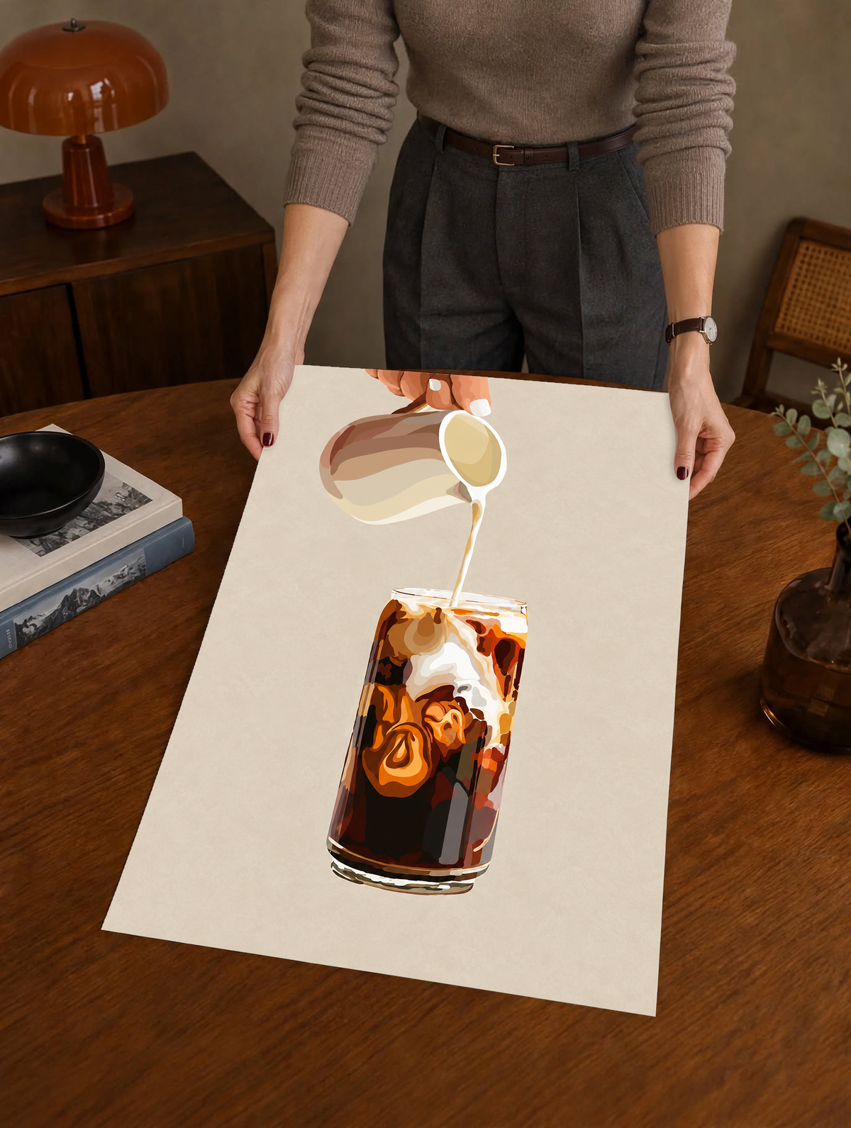 Mockup eines ungerahmten Drucks, von einer Person in einem warmen Innenraum gehalten: Eiskaffeeillustration mit einer Hand, die Milch aus einem gestreiften Kännchen in ein hohes Glas gießt, und cremefarbenen Karamellwirbeln auf Beige.