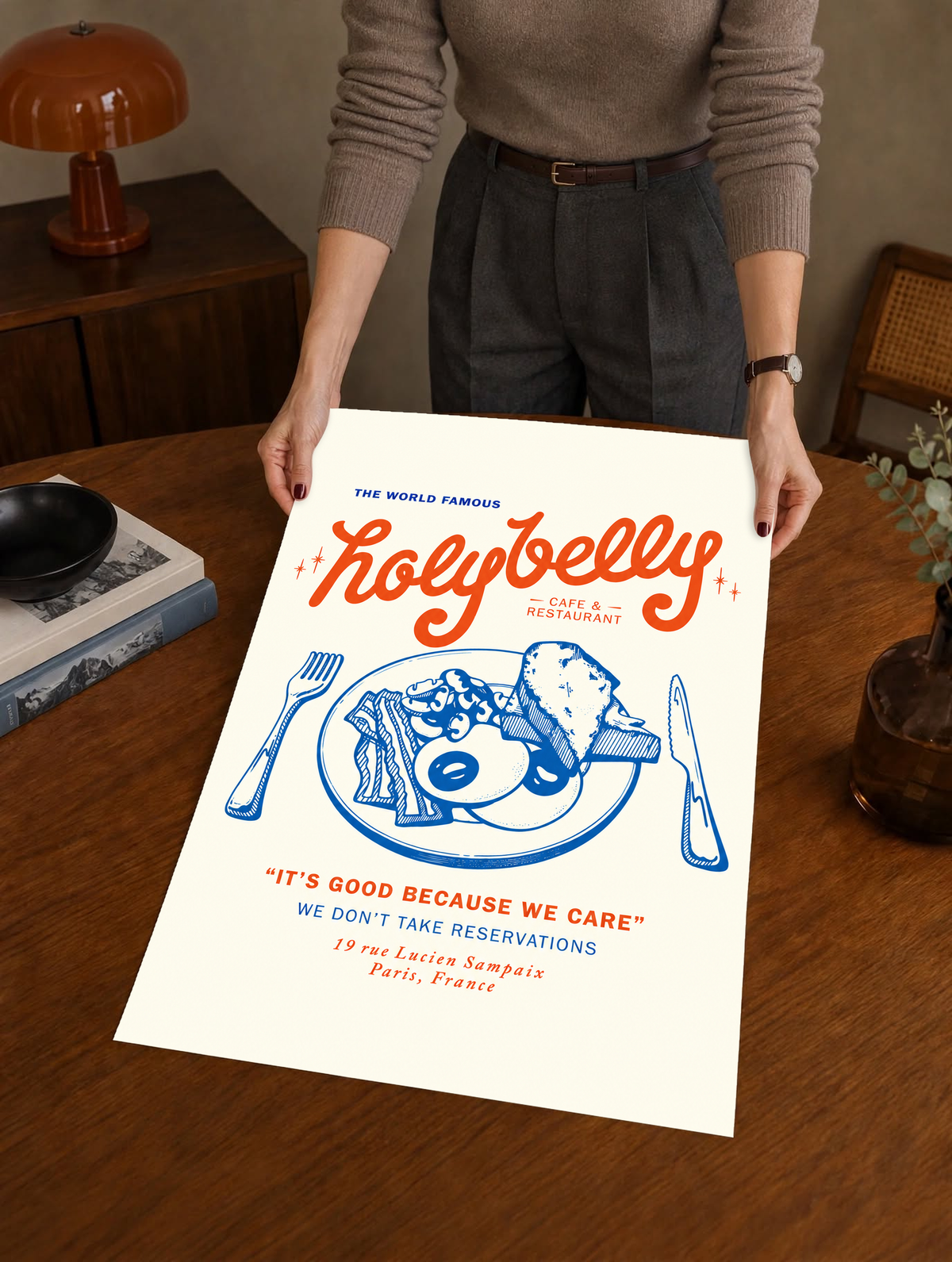 Ambientazione della stampa senza cornice tenuta da una persona in un interno caldo: Illustrazione della colazione Holybelly con grandi scritte rosse, piatto blu con uova e pane tostato, forchetta, coltello e testi da caffetteria su crema.