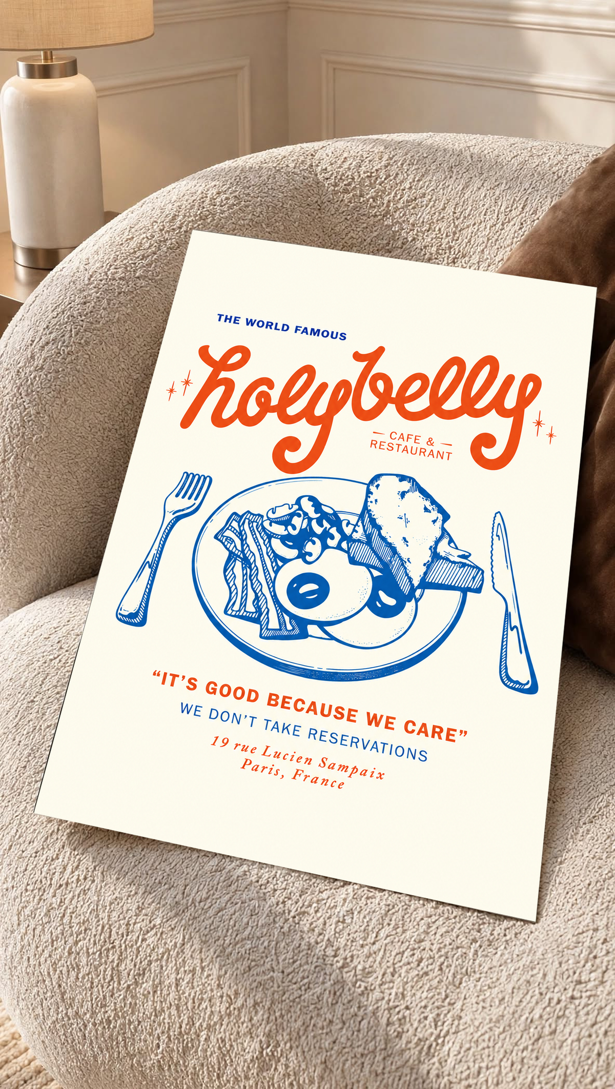 Ambientazione della stampa senza cornice su una poltrona crema dal tessuto strutturato: Illustrazione della colazione Holybelly con grandi scritte rosse, piatto blu con uova e pane tostato, forchetta, coltello e testi da caffetteria su crema.