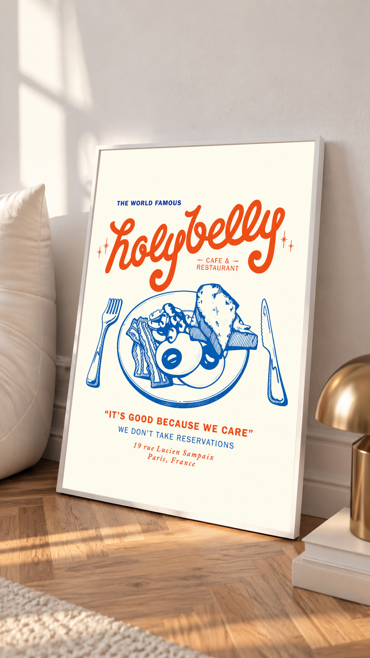 Ambientazione in cornice bianca appoggiata a una parete chiara accanto a un cuscino: Illustrazione della colazione Holybelly con grandi scritte rosse, piatto blu con uova e pane tostato, forchetta, coltello e testi da caffetteria su crema.
