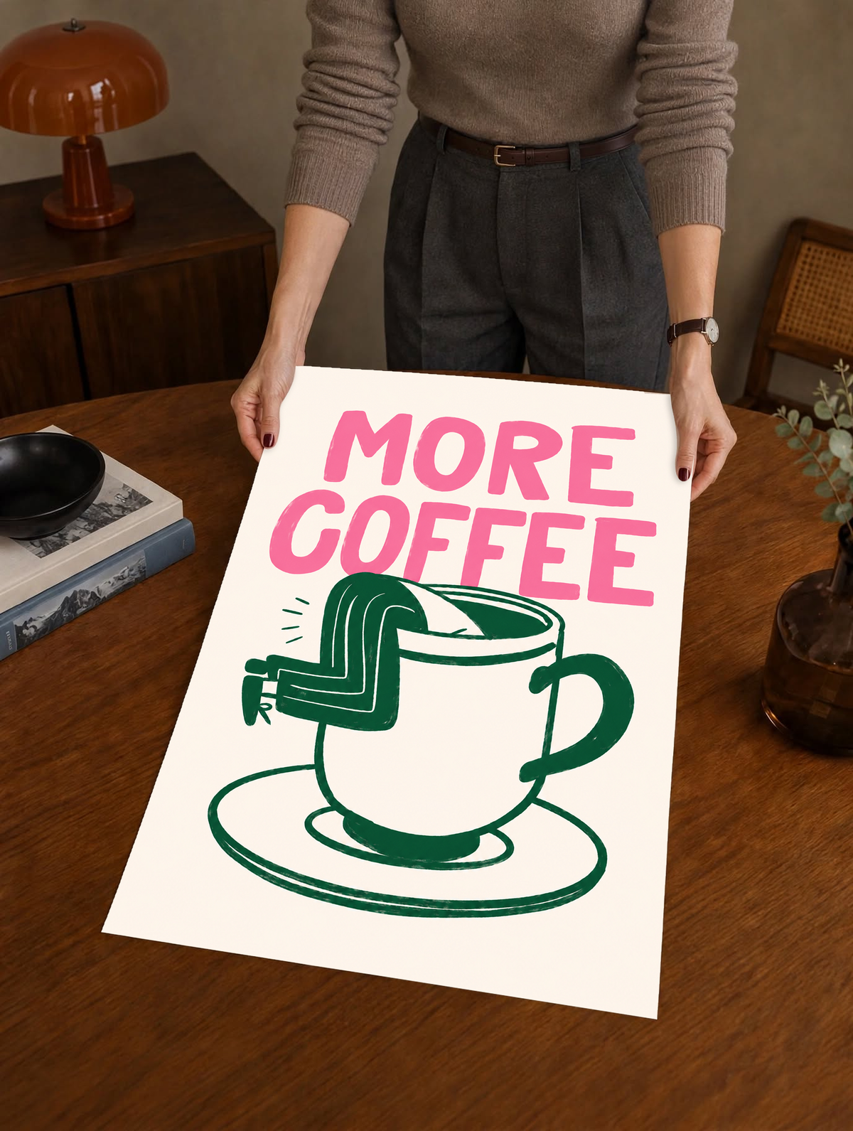Mockup eines ungerahmten Drucks, von einer Person in einem warmen Innenraum gehalten: Großes rosa MORE COFFEE über einer grün gezeichneten Tasse mit Untertasse und einem Beinpaar über dem Rand auf Creme.