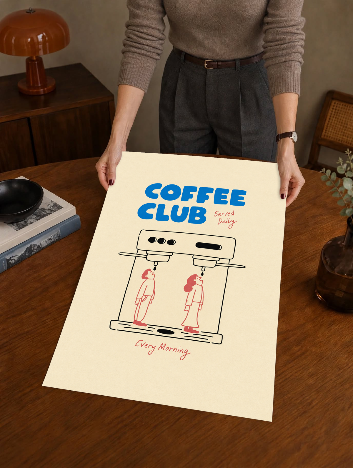 Blaues Coffee Club-Motiv mit schwarz gezeichneter Espressomaschine, zwei korallroten Figuren, roter Schrift und cremefarbenem Hintergrund. Ungerahmtes Poster, über einem Holztisch neben einem Plattenspieler gehalten.