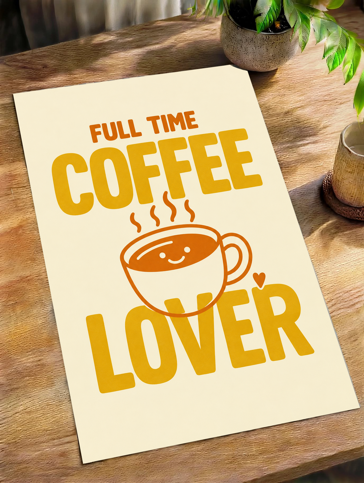 Full Time Coffee Lover-Motiv mit goldgelber Schrift, orangefarbener lächelnder Kaffeetasse, Dampf und kleinem Herz auf Creme. Ungerahmtes Poster schräg auf einem Holztisch neben Pflanze und Tasse.