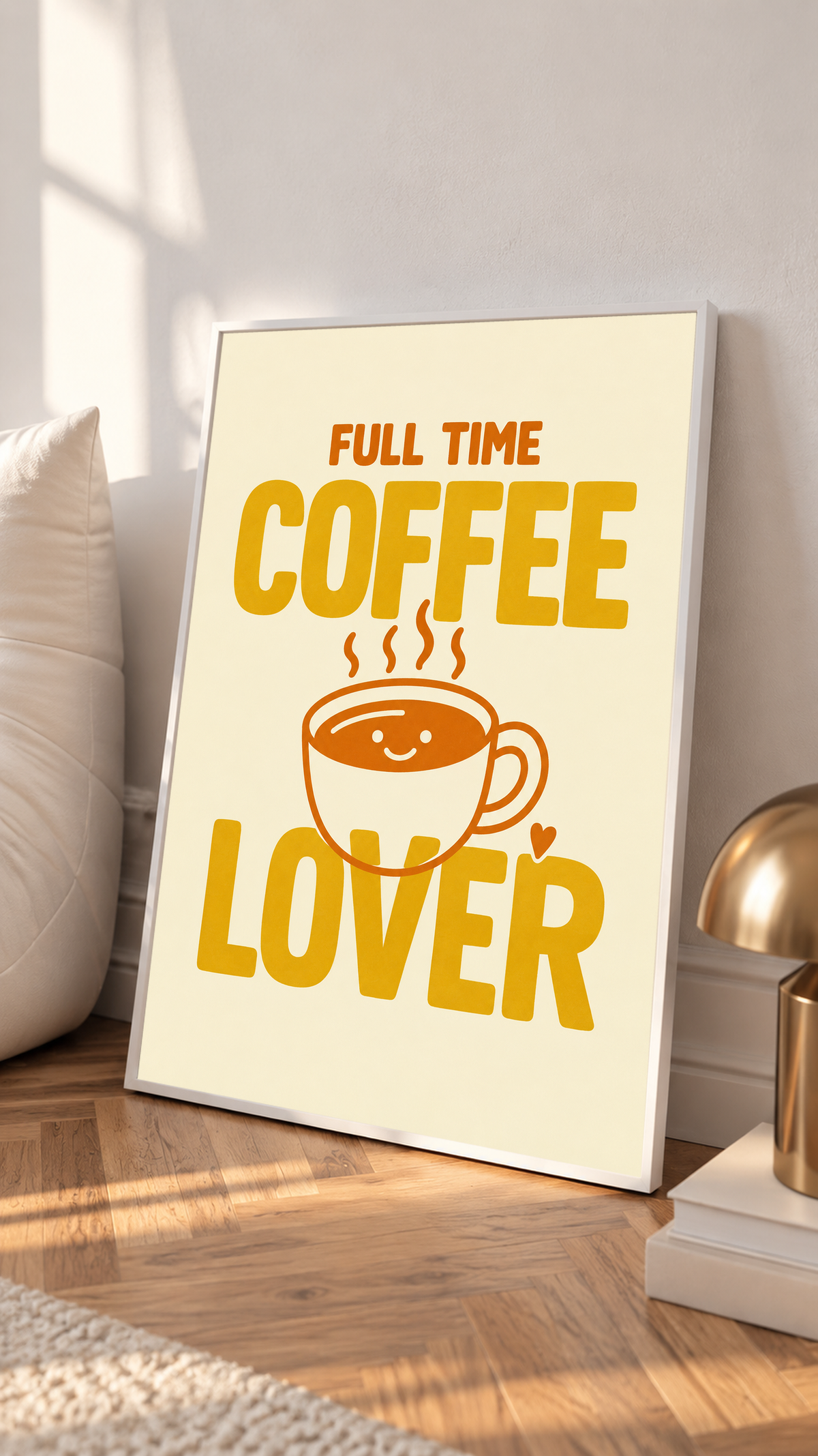 Full Time Coffee Lover-Motiv mit goldgelber Schrift, orangefarbener lächelnder Kaffeetasse, Dampf und kleinem Herz auf Creme. Im weißen Rahmen auf einem Holzboden, neben Kissen und einer kleinen Leuchte.