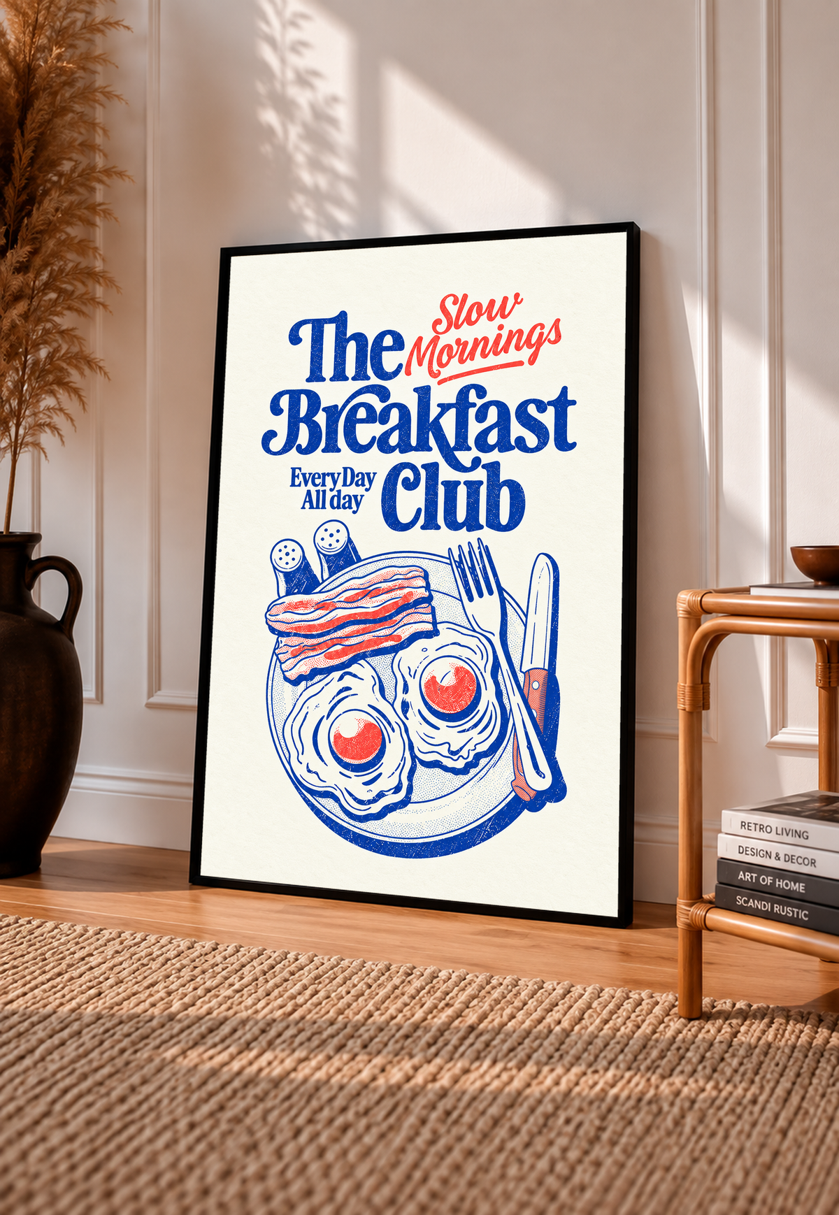 Slow Mornings Breakfast Club-Motiv mit blauer Schrift, roter Schreibschrift, zwei Spiegeleiern, Speck, Besteck und Streuern auf Creme. Im schwarzen Rahmen an einer sonnigen Wand, neben Holzstuhl und gewebtem Teppich.