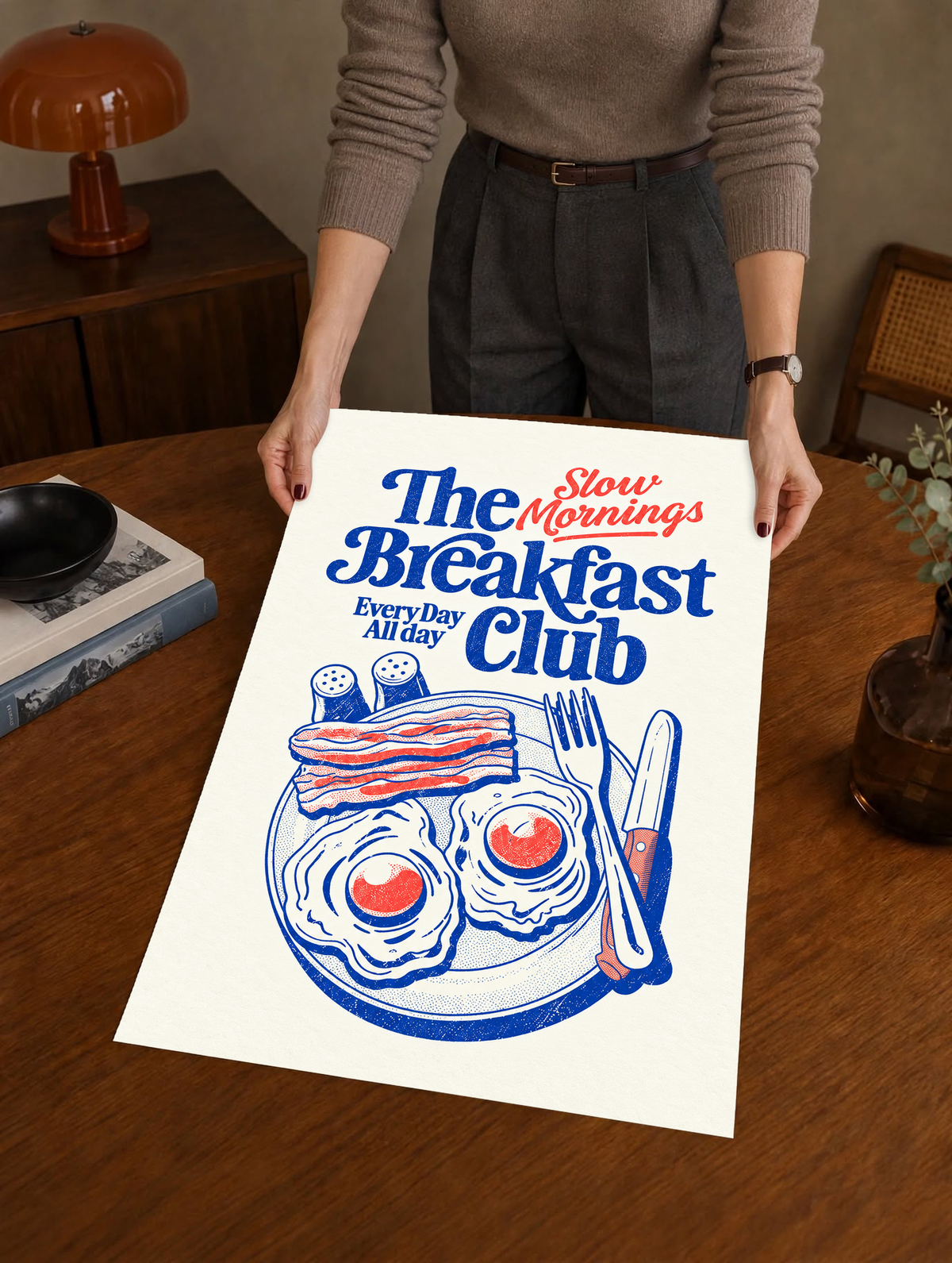 Slow Mornings Breakfast Club-Motiv mit blauer Schrift, roter Schreibschrift, zwei Spiegeleiern, Speck, Besteck und Streuern auf Creme. Ungerahmtes Poster, über einem Holztisch neben einem Plattenspieler gehalten.