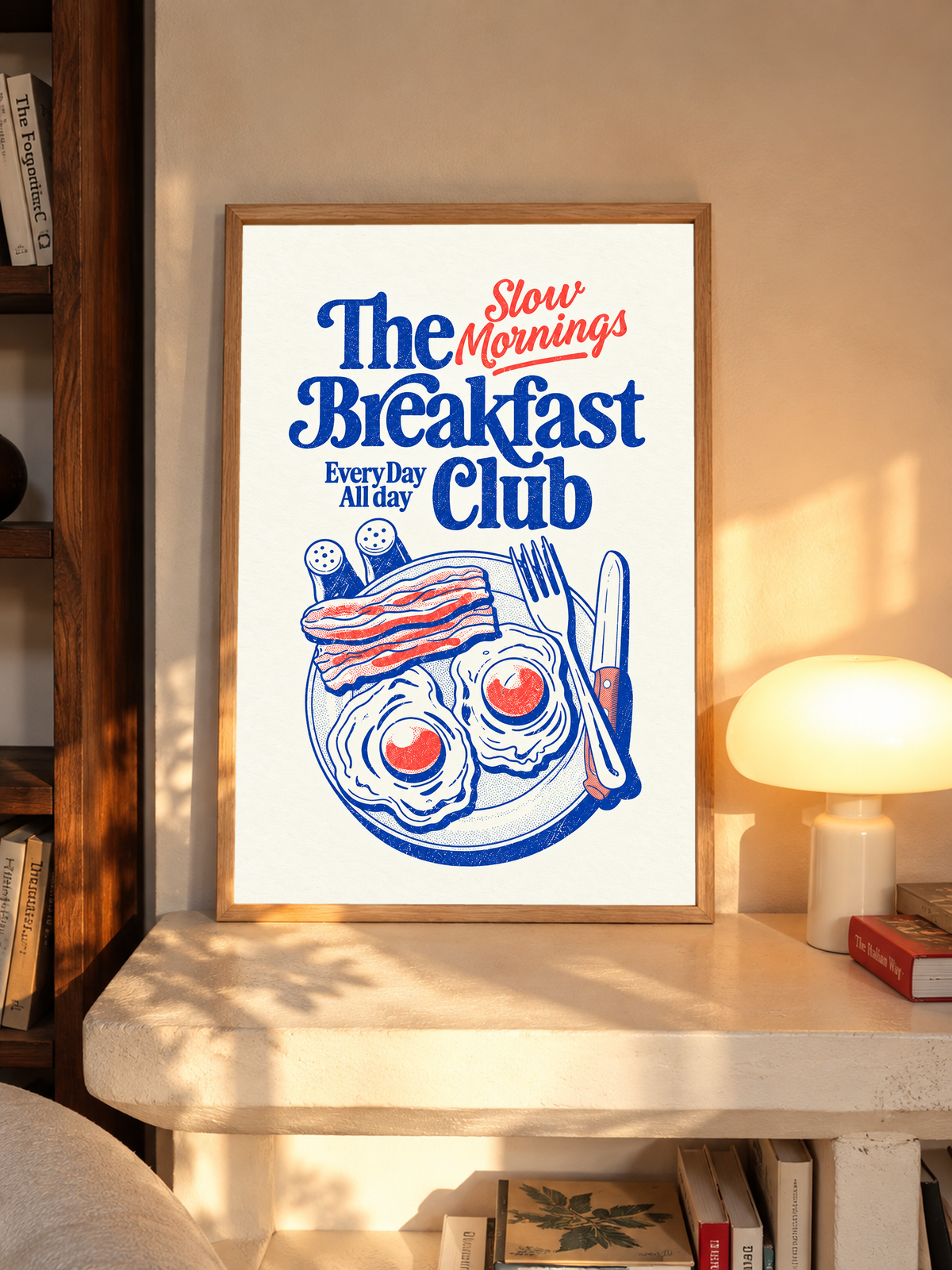 Slow Mornings Breakfast Club-Motiv mit blauer Schrift, roter Schreibschrift, zwei Spiegeleiern, Speck, Besteck und Streuern auf Creme. Im Eichenrahmen auf einem Schreibtisch neben einer leuchtenden Pilzleuchte.