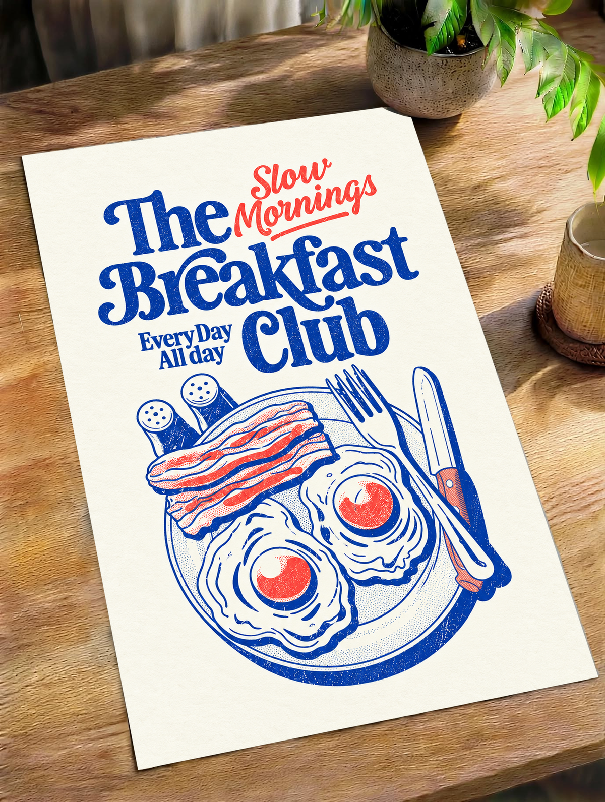 Slow Mornings Breakfast Club-Motiv mit blauer Schrift, roter Schreibschrift, zwei Spiegeleiern, Speck, Besteck und Streuern auf Creme. Ungerahmtes Poster schräg auf einem Holztisch neben Pflanze und Tasse.