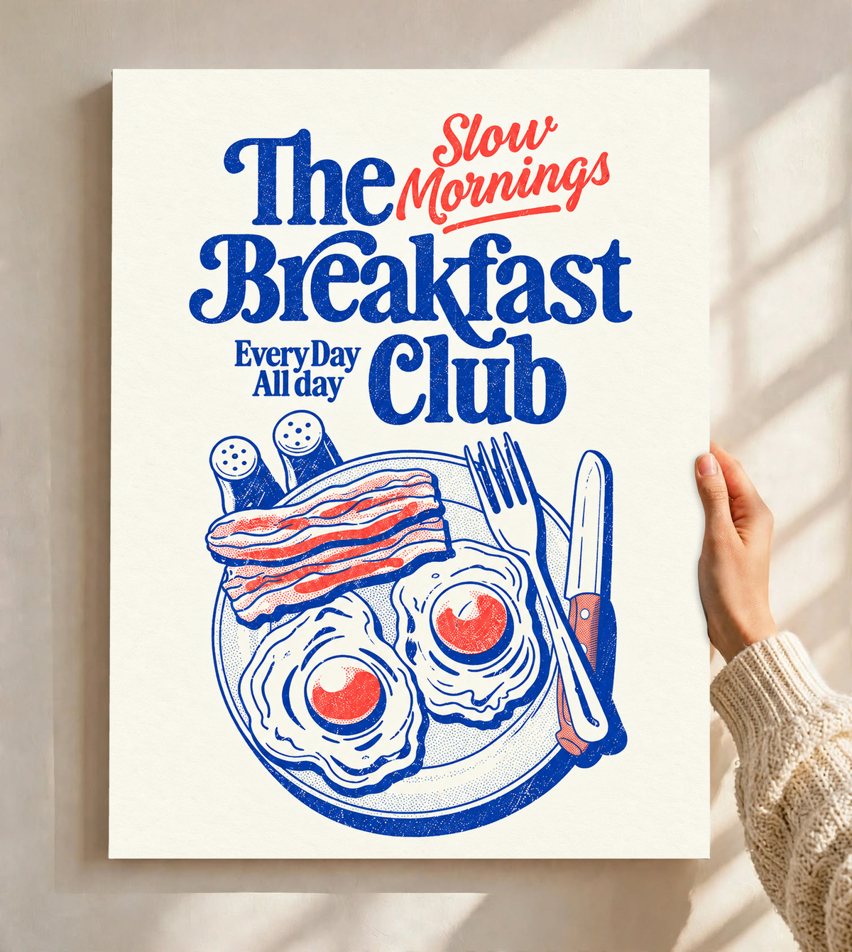 Slow Mornings Breakfast Club-Motiv mit blauer Schrift, roter Schreibschrift, zwei Spiegeleiern, Speck, Besteck und Streuern auf Creme. Ungerahmtes Poster, vor einer hellen Wand mit Fensterschatten gehalten.