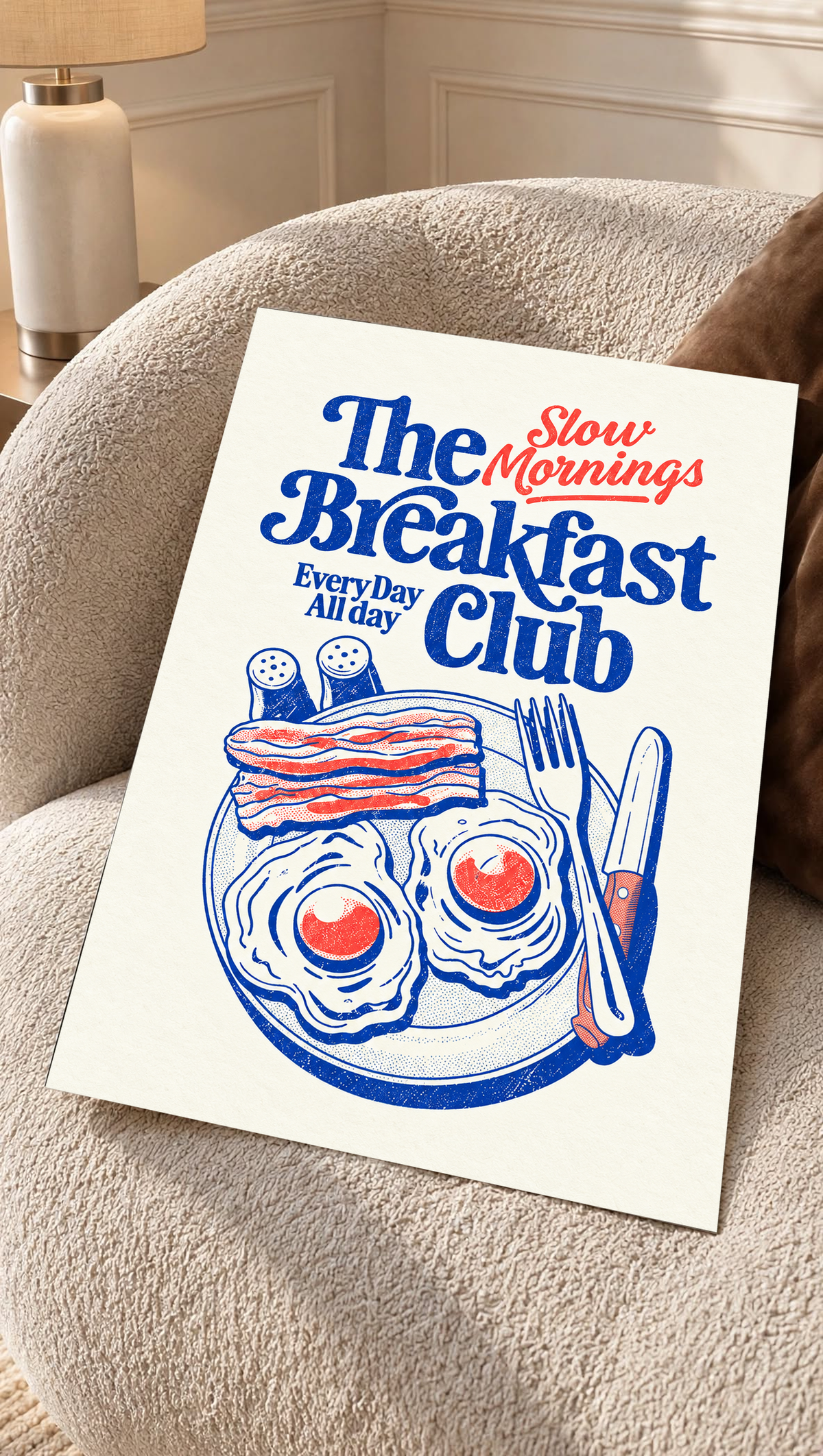Slow Mornings Breakfast Club-Motiv mit blauer Schrift, roter Schreibschrift, zwei Spiegeleiern, Speck, Besteck und Streuern auf Creme. Ungerahmtes Poster auf einem strukturierten cremefarbenen Sessel.